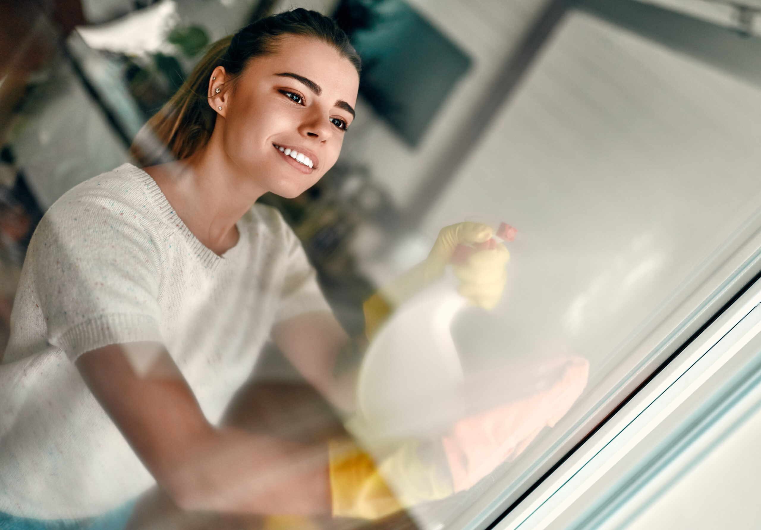 A young attractive woman in rubber gloves and casual home clothes washes a glass with a special spray and rag.