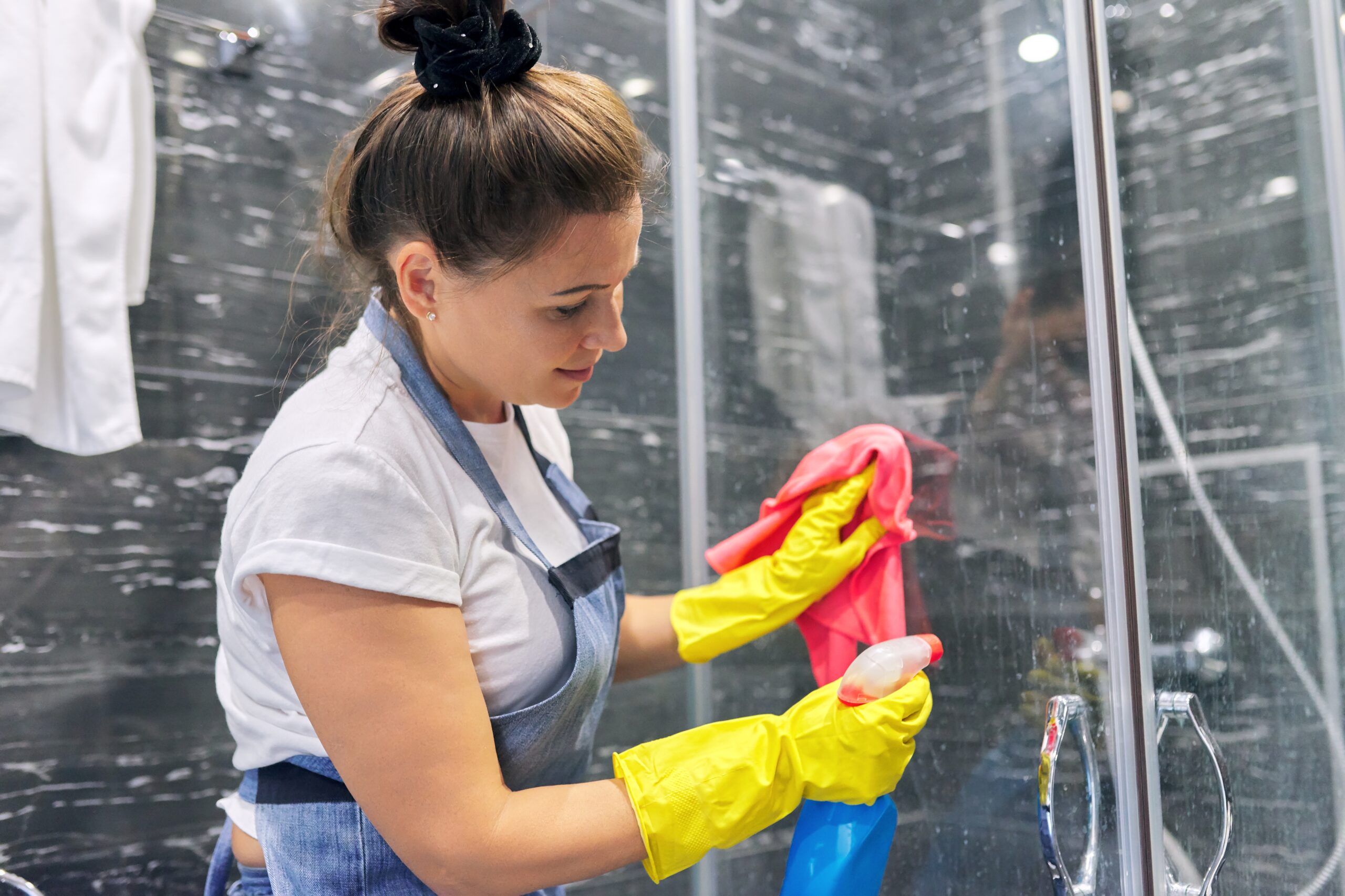 Cleaning lady in apron gloves with detergent washing glass of shower cabin in bathroom, house cleaning